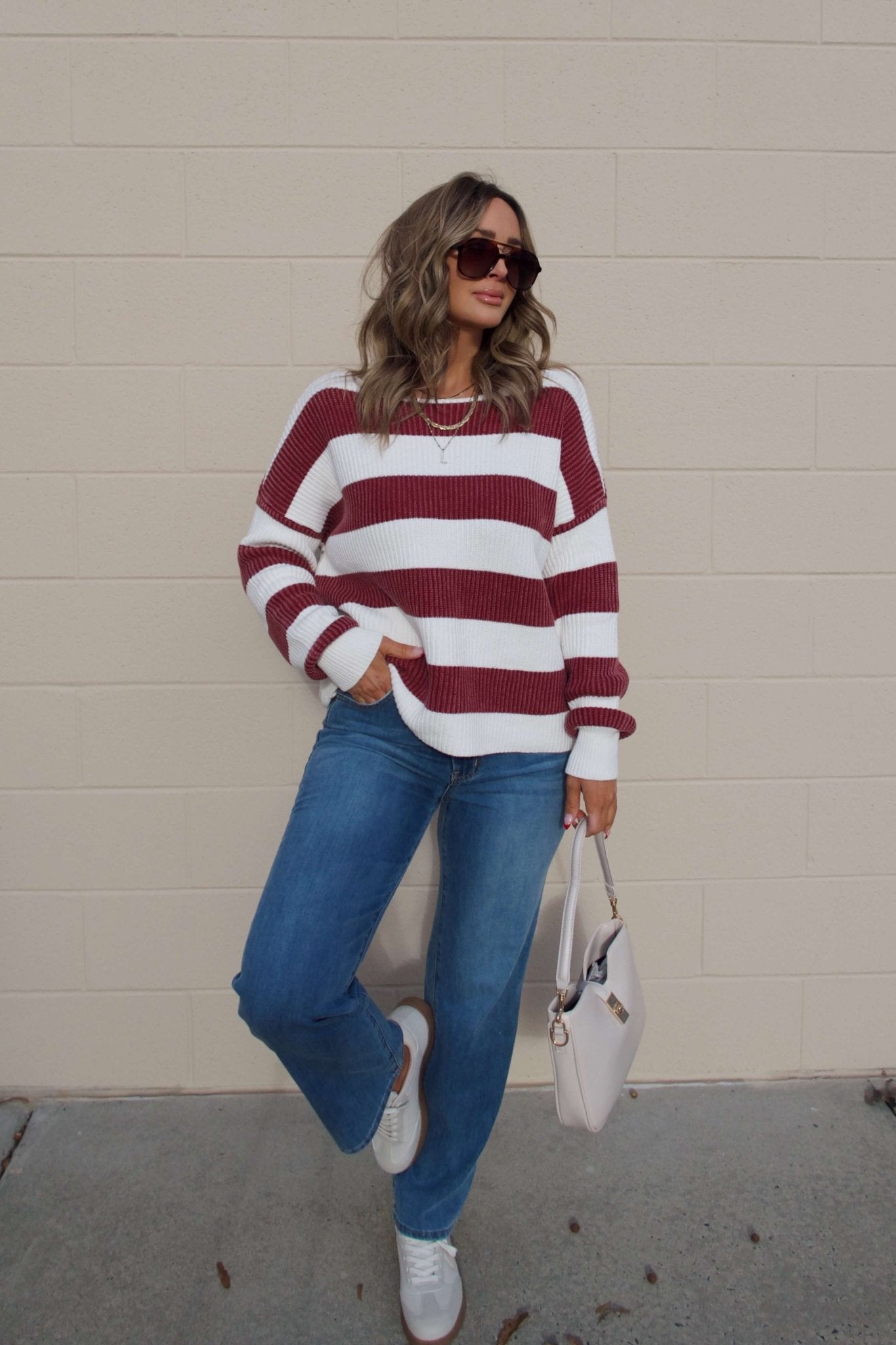 Woman wearing a Callahan Wine and White Striped Sweater, blue jeans, sunglasses, and holding a white handbag by a beige wall.