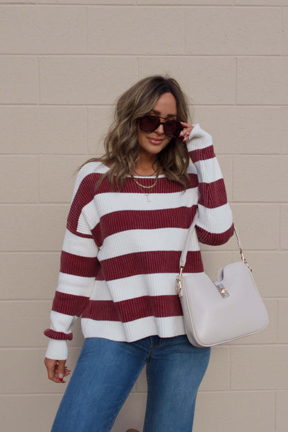 Woman in sunglasses, blue jeans, and a Callahan Wine and White Striped Sweater holds a beige handbag by a tan brick wall.