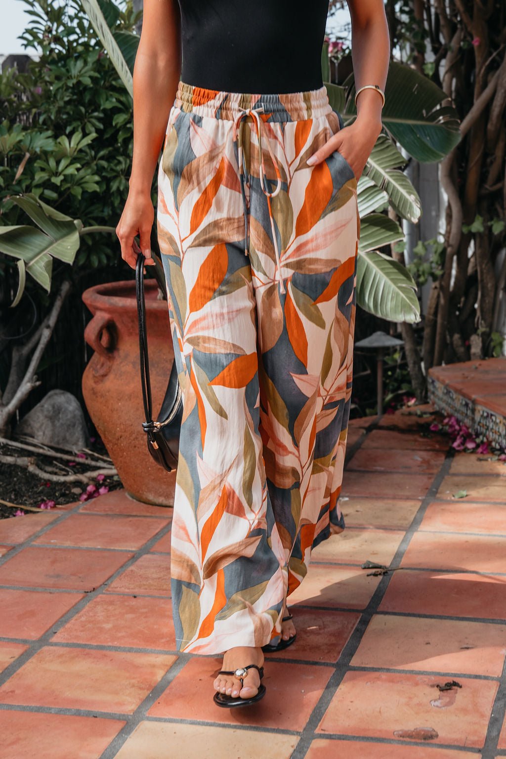 Wearing Calypso Multi Leaf Wide Leg Pants in orange, green, and beige, a person walks on a tiled patio.
