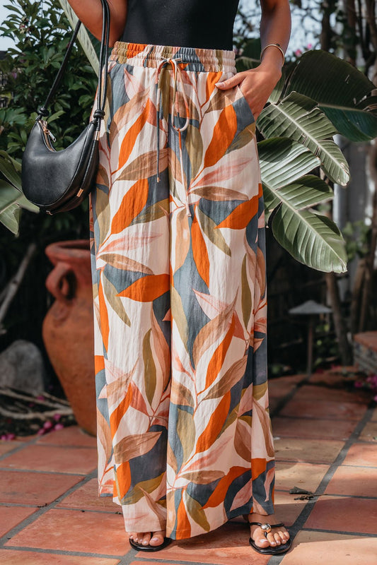 A person in Calypso Multi Leaf Wide Leg Pants, black sandals, and a black top stands on a tiled patio.
