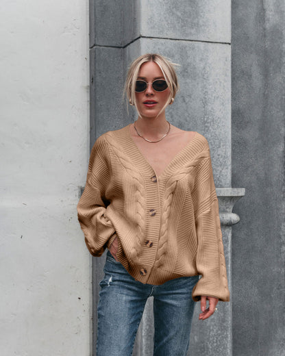 A woman in sunglasses, blue jeans, and the Camden Button Down Cardigan - Taupe stands against a gray and white stone wall.