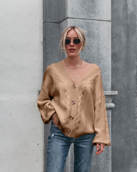 A woman in sunglasses wears the Camden Button Down Cardigan - Taupe with blue jeans, standing by a textured gray and white wall.