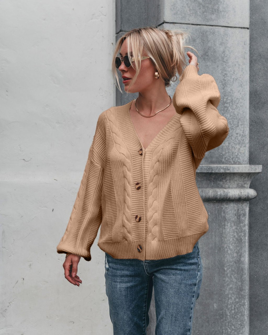 Woman in taupe Camden Button Down Cardigan – DOORBUSTER, sunglasses, and blue jeans adjusts her hair by a gray and white wall.