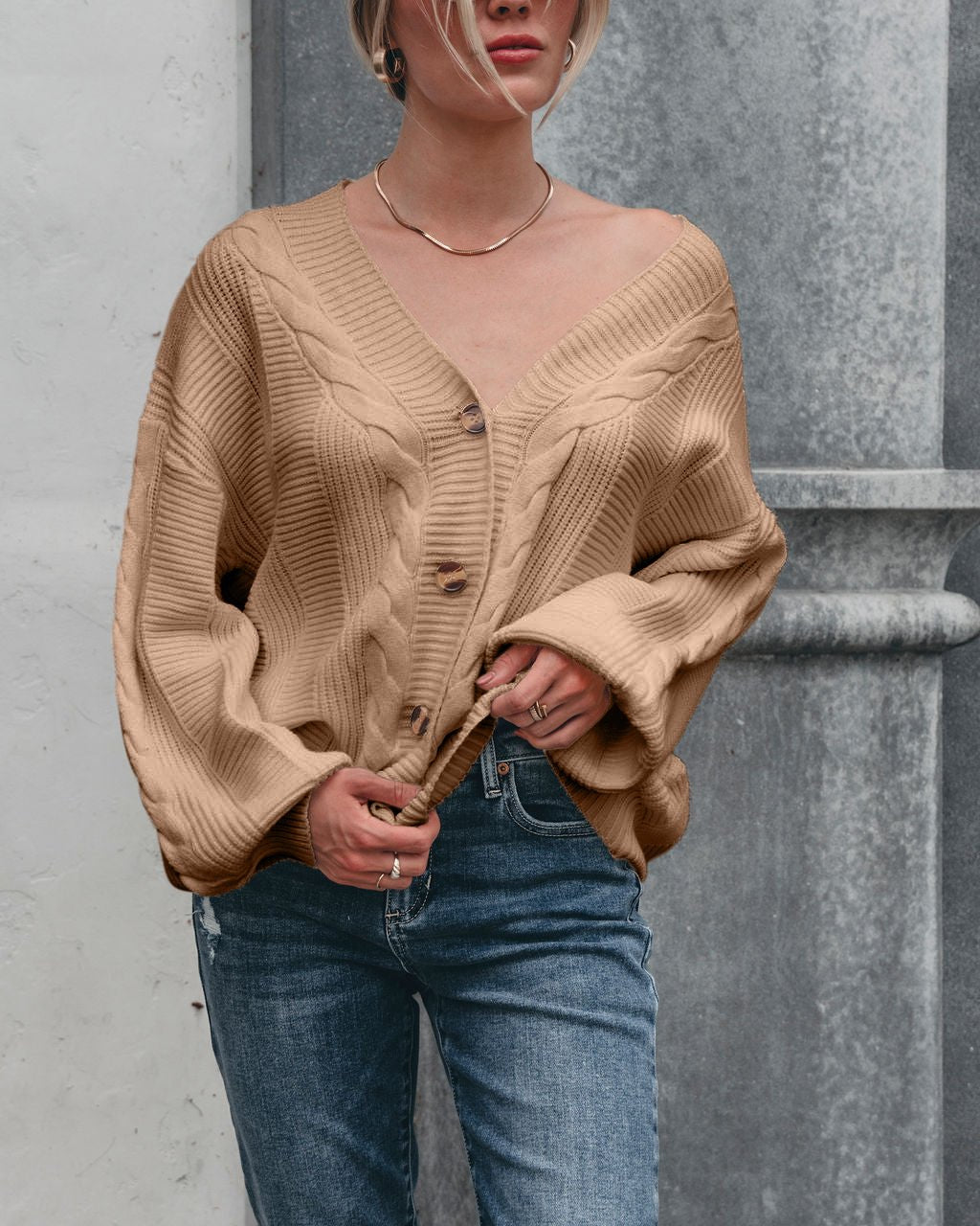 A woman in the Camden Button Down Cardigan - Taupe poses in blue jeans against a gray stone background.