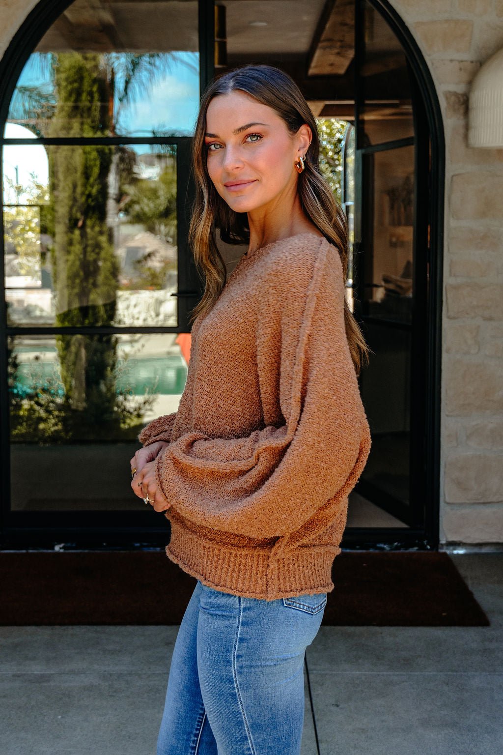 A woman in a Camel Boat Neck Seam Detail Sweater and blue jeans stands sideways outdoors, smiling by large glass doors.