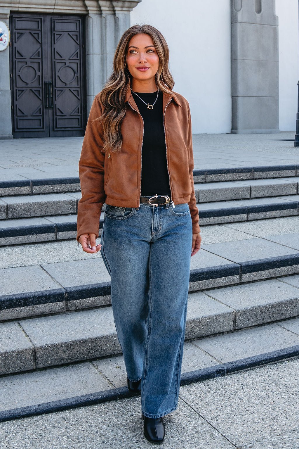 Woman with long wavy hair in a Camel Suede Zip Up Jacket, black shirt, and wide-leg jeans walks down outdoor steps.