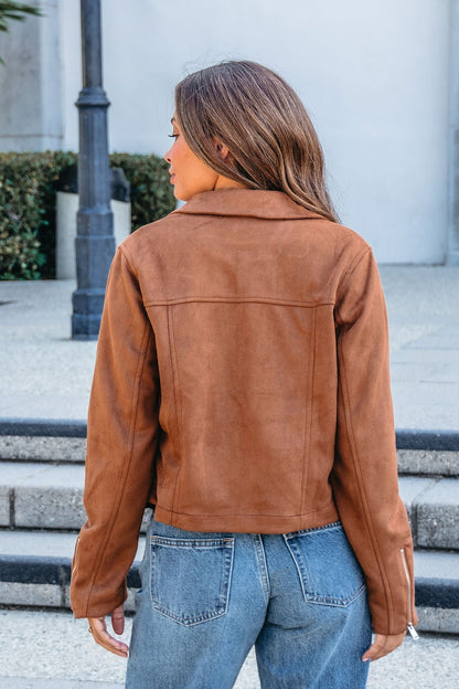 A woman with long brown hair models the Camel Suede Zip Up Jacket and blue jeans outdoors, seen from behind.