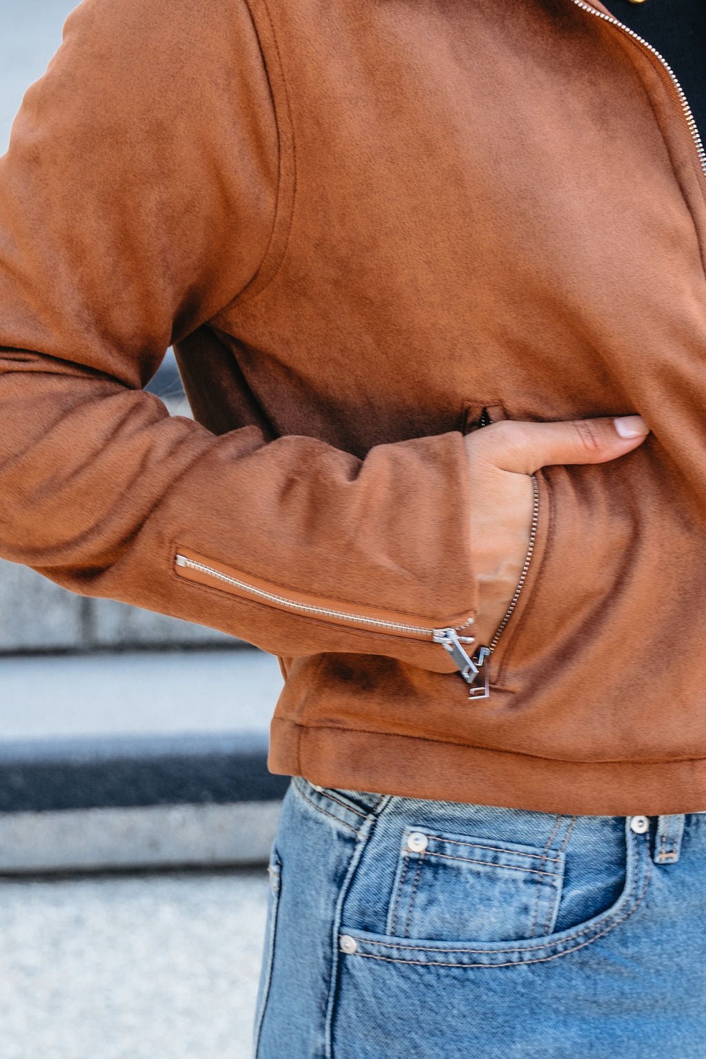 Someone wearing a Camel Suede Zip Up Jacket and blue jeans stands with one hand in their pocket.