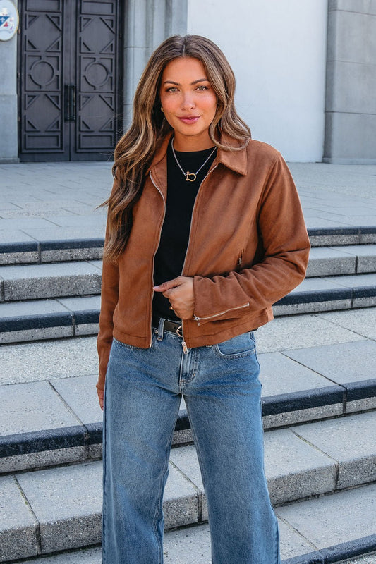 Woman with long brown hair wears a Camel Suede Zip Up Jacket, black top, and blue jeans in front of stone steps and a large door.