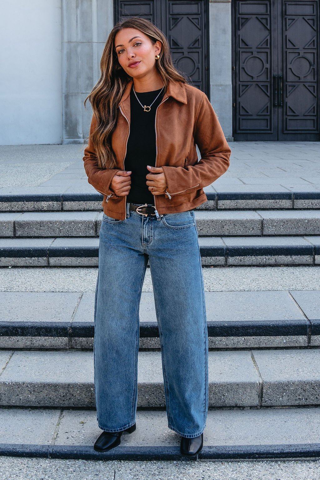 Woman poses confidently on steps in a Camel Suede Zip Up Jacket, black top, wide-leg jeans, and black shoes.