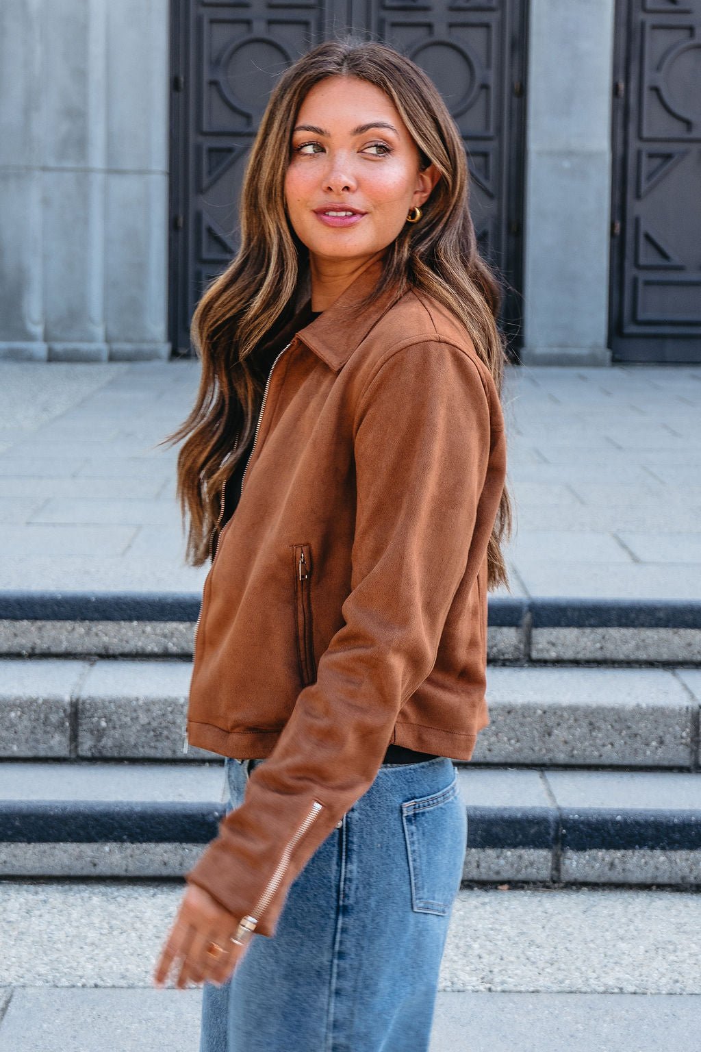 Wearing the Camel Suede Zip Up Jacket and blue jeans, a woman stands outdoors, gazing to the side with a slight smile.