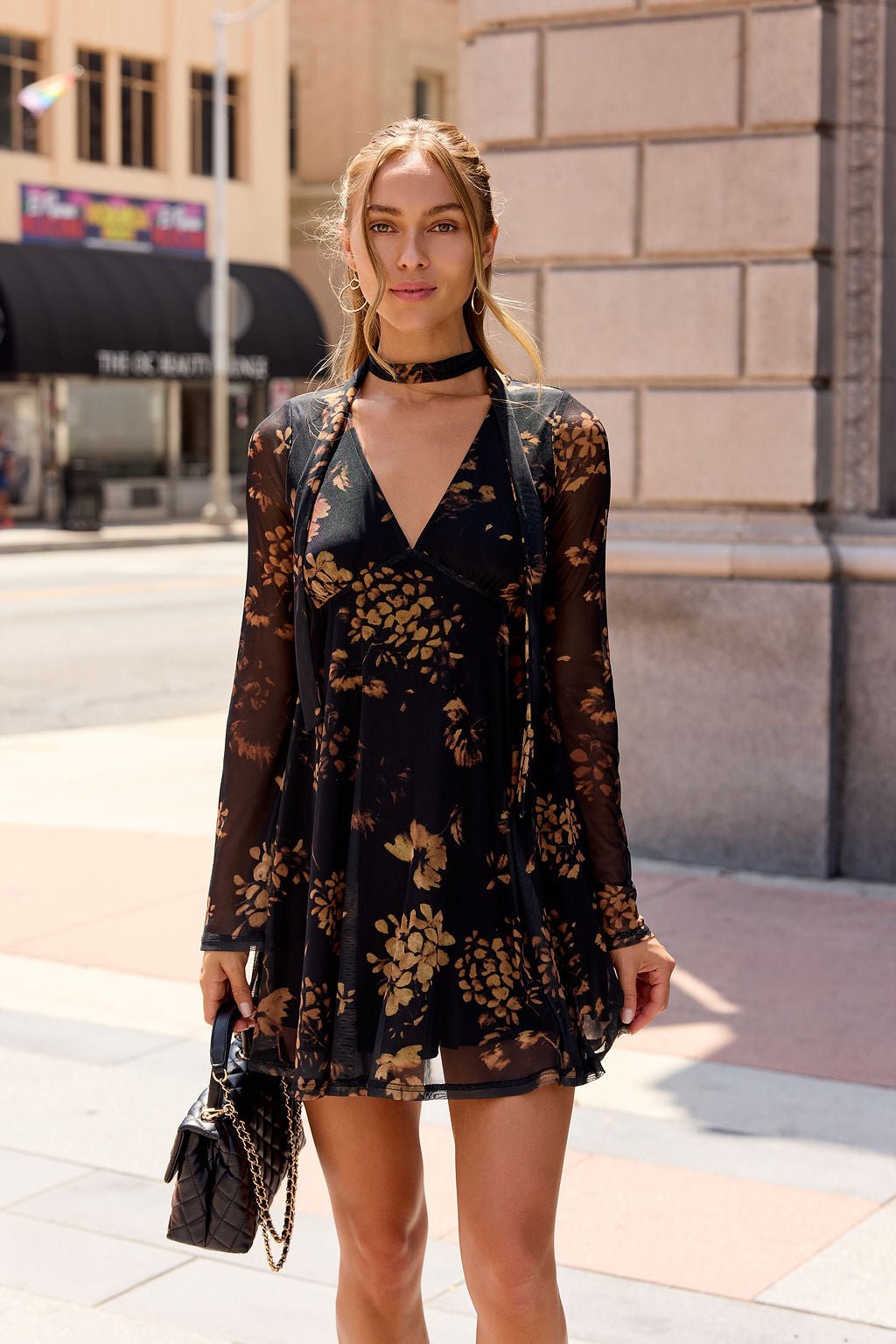 Woman wears the Camellia Floral Mesh Mini Dress - Black, long-sleeved, holding a black purse on a sunny city sidewalk.
