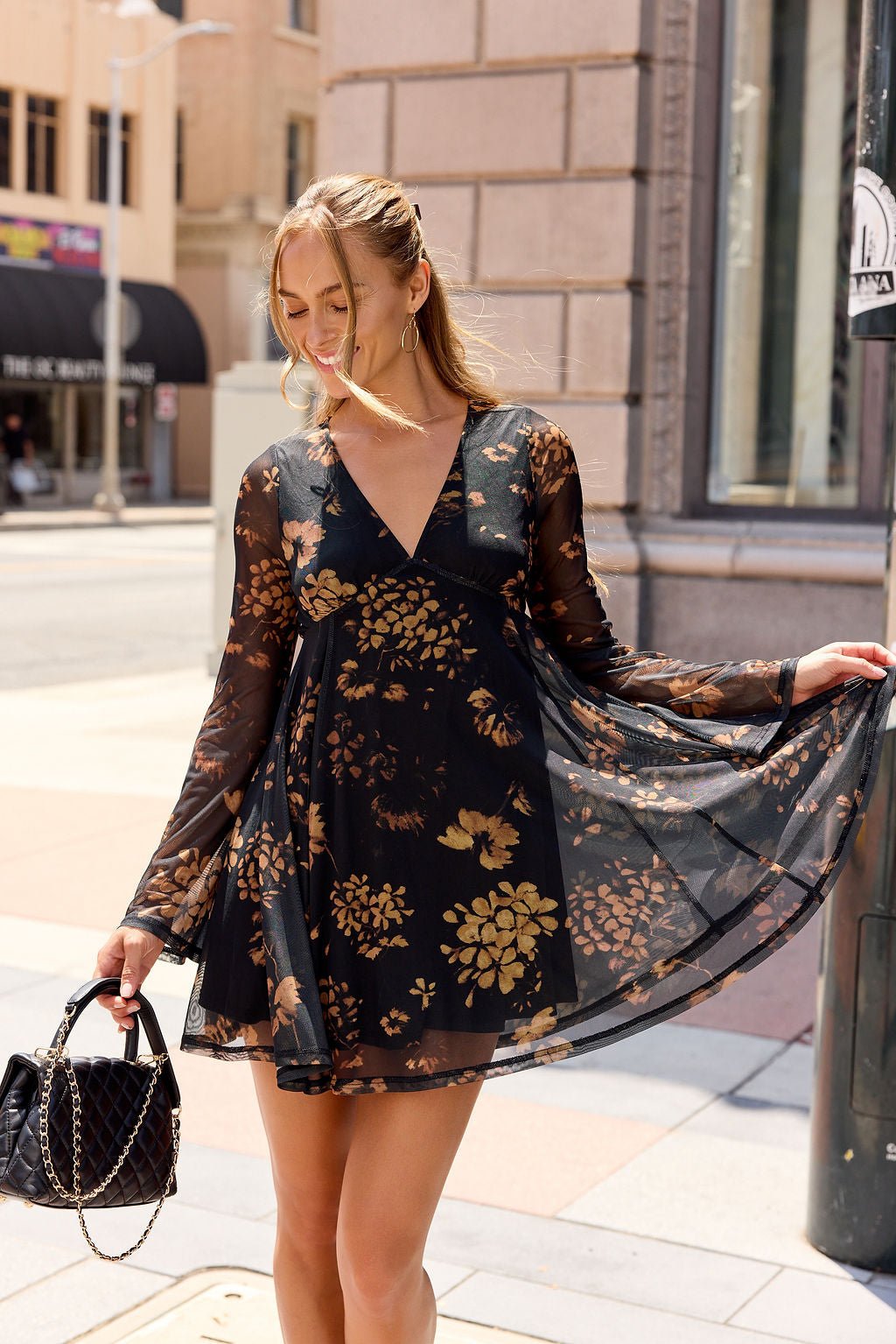 Woman in the Camellia Floral Mesh Mini Dress - Black stands on a city sidewalk, holding a black purse and smiling.