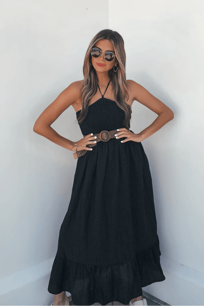 Woman wears the Camille Black Halter Belted Midi Dress, sunglasses, and hoop earrings, standing against a white wall.