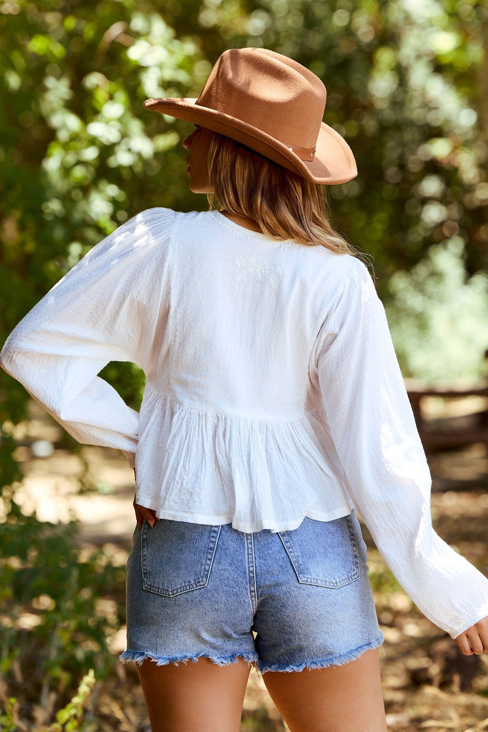 A person in the Candace White Embroidered Peplum Top - FINAL SALE and denim shorts stands outdoors, facing away.