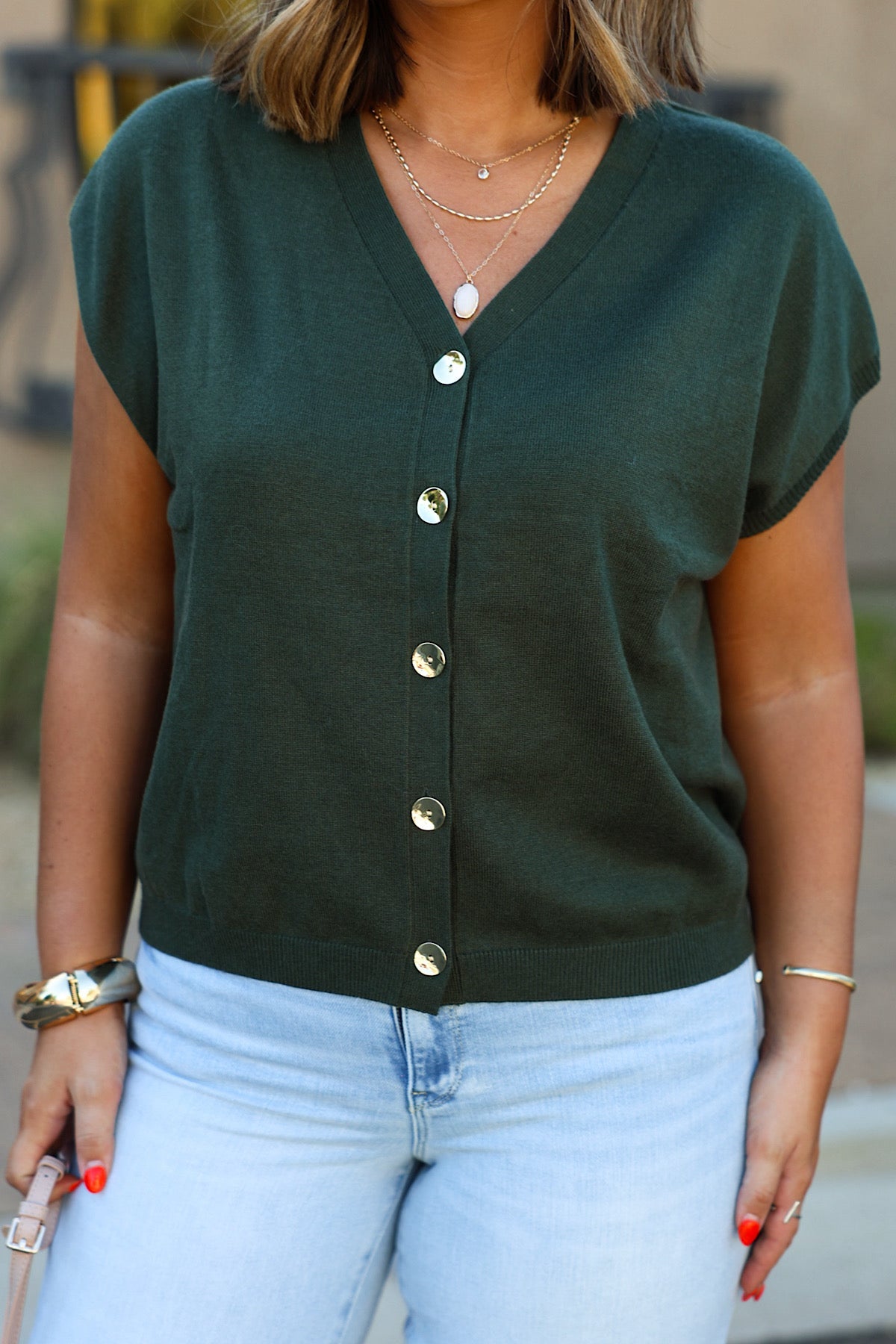 Woman in an olive cap sleeve button down sweater, layered necklaces, and light jeans holds a pink item outdoors.