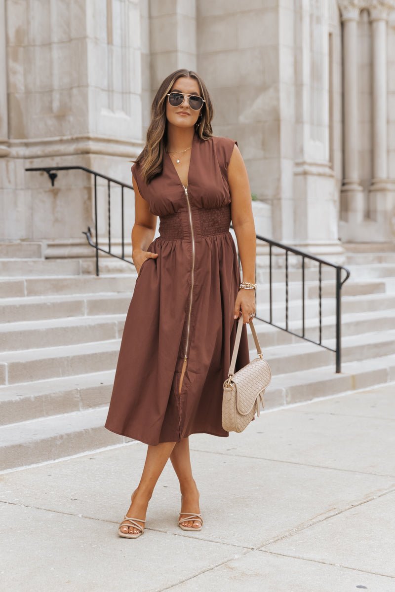 Woman in the Cap Sleeve Smocked Front Zip Shirt Dress in brown with sunglasses, sandals, and a beige handbag on stone steps.