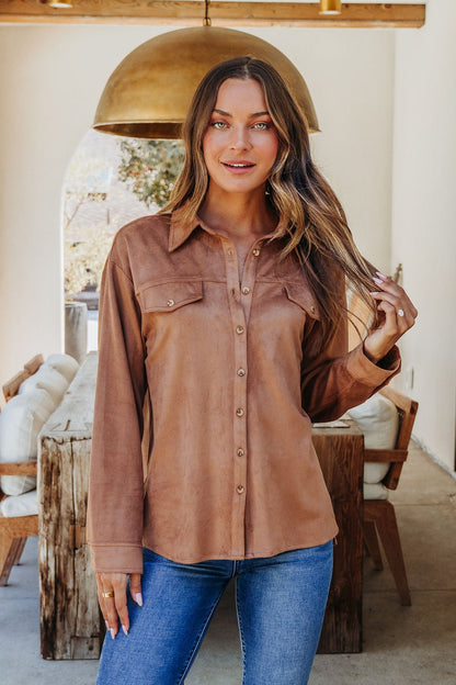 A woman wearing a Carmel Brown Faux Suede Shacket and blue jeans stands indoors beside rustic wooden furniture, touching her hair.