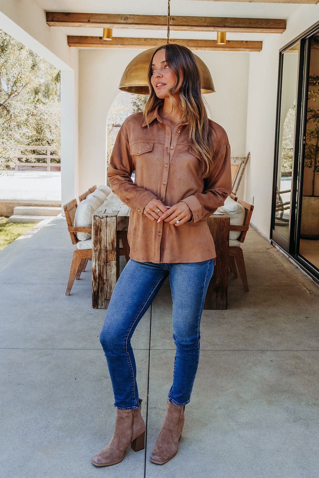 A woman in a Carmel Brown Faux Suede Shacket, blue jeans, and boots stands by a wooden dining table, looking to the side.