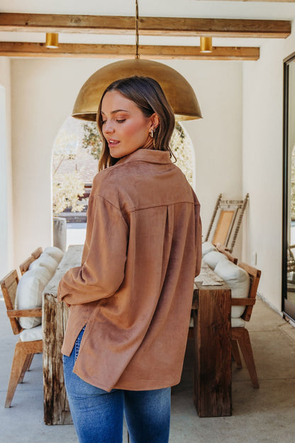 A woman in a Carmel Brown Faux Suede Shacket and blue jeans stands indoors in a chic dining area with wooden furniture.