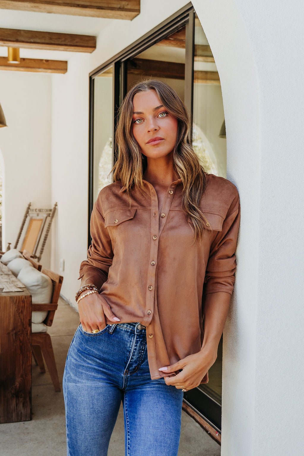 A woman in a Carmel Brown Faux Suede Shacket and blue jeans stands outside by a white wall, looking at the camera.