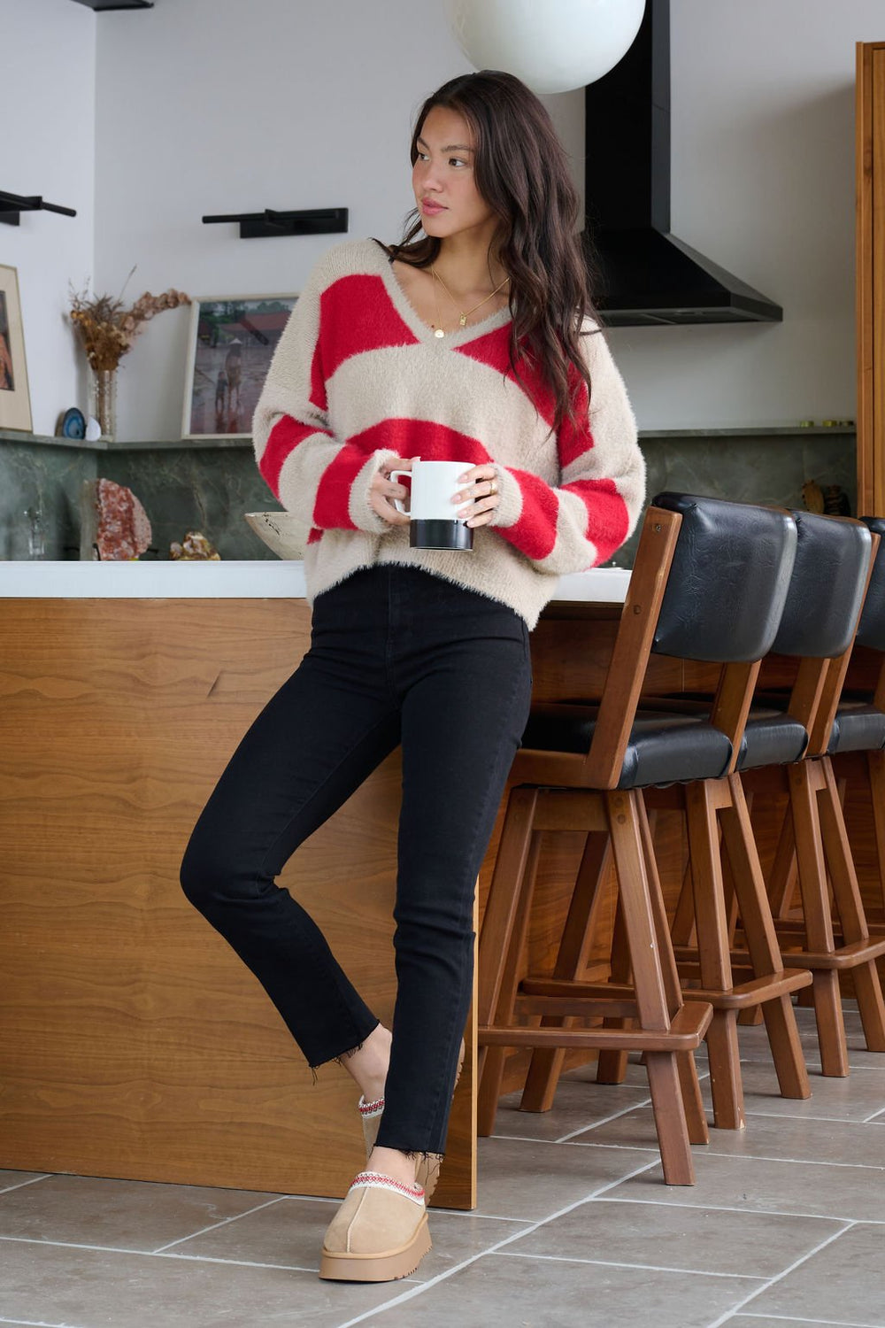 Woman in the Carmen Red and Taupe Striped Sweater stands by a kitchen counter holding a mug.