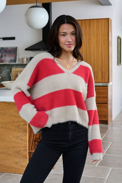 A woman wears the Carmen Red and Taupe Striped Sweater with black pants, smiling in a modern kitchen.