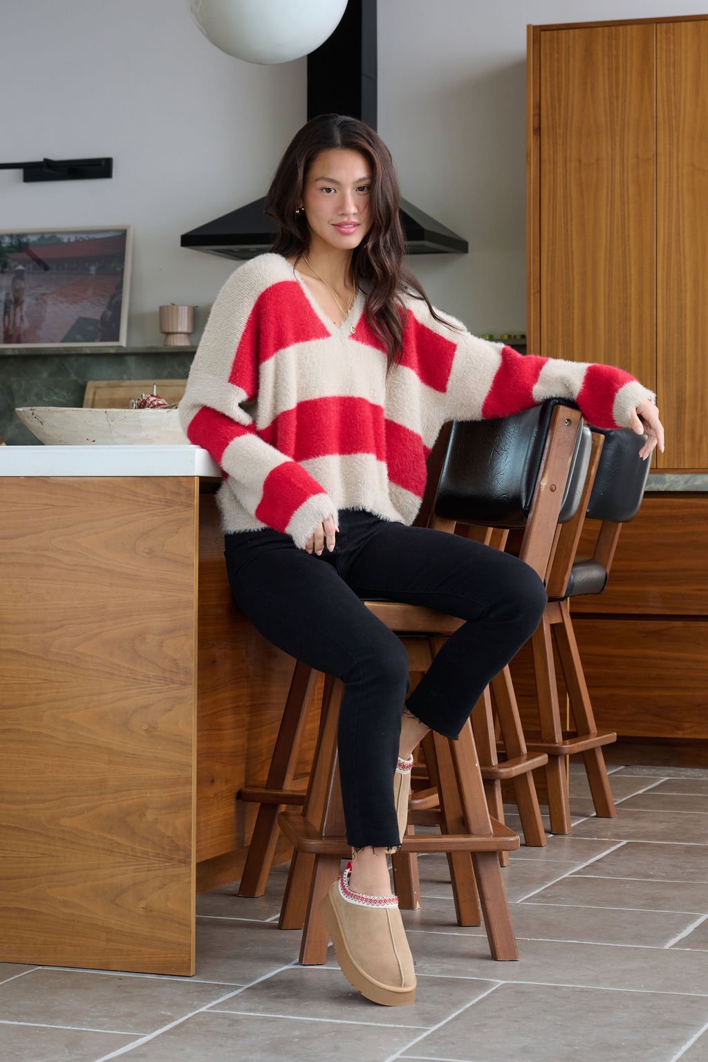 A woman in the Carmen Red and Taupe Striped Sweater sits on a barstool in a modern kitchen, wearing black pants and tan slippers.