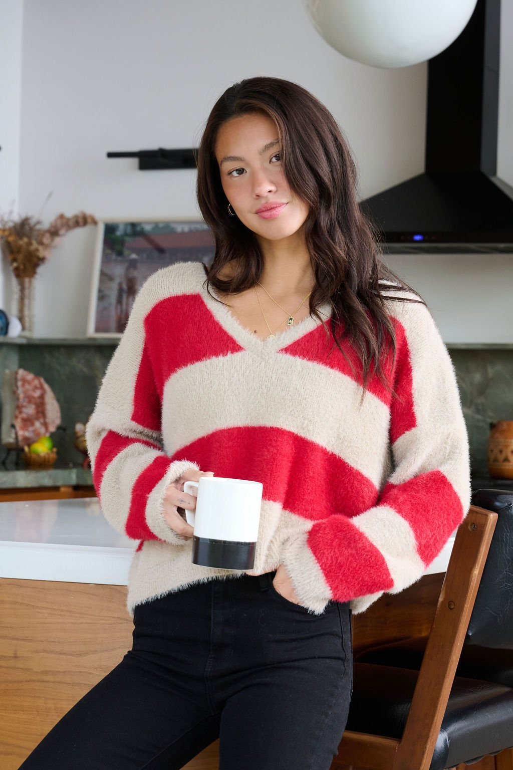 Woman with long dark hair wears the Carmen Red and Taupe Striped Sweater in a modern kitchen, holding a mug and looking at the camera.