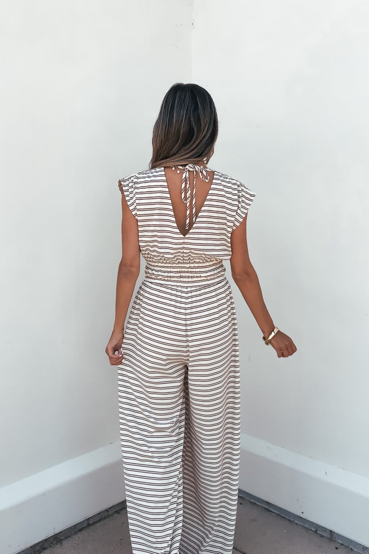 Woman in the Carrie Stripe Wide Leg Jumpsuit - Brown/Cream, seen from the back, stands against a plain white wall.