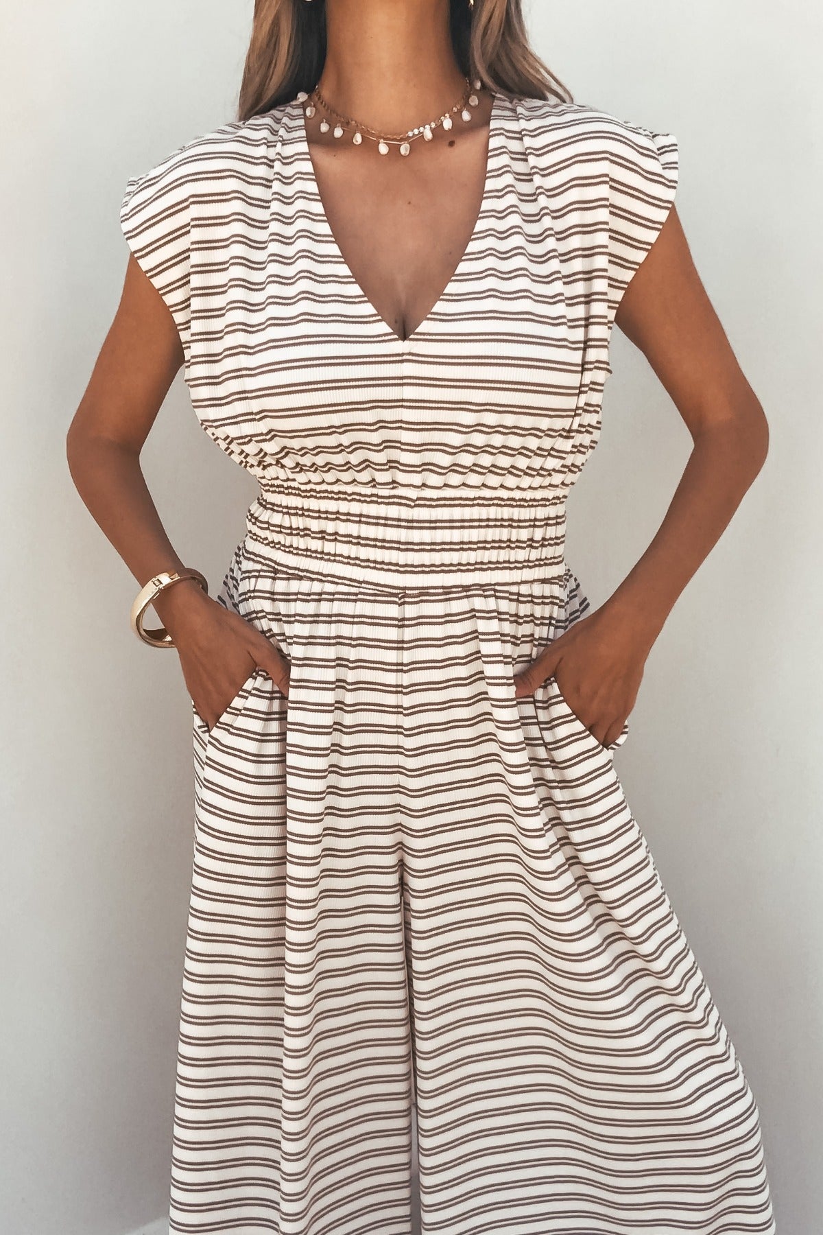 Woman in the Carrie Stripe Wide Leg Jumpsuit - Brown/Cream, accessorized with a necklace and bracelet, hands in pockets.