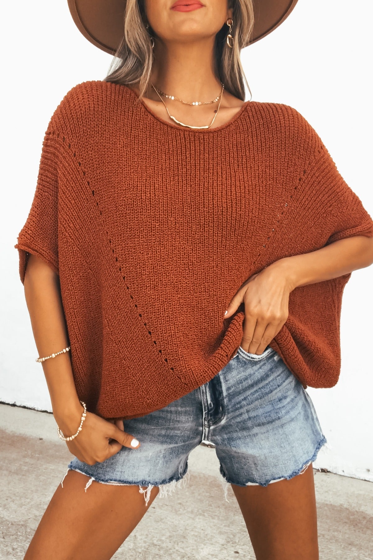 Woman in the Cassia Boatneck Sweater - Rust, denim shorts, brown hat, and jewelry, standing against a white background.
