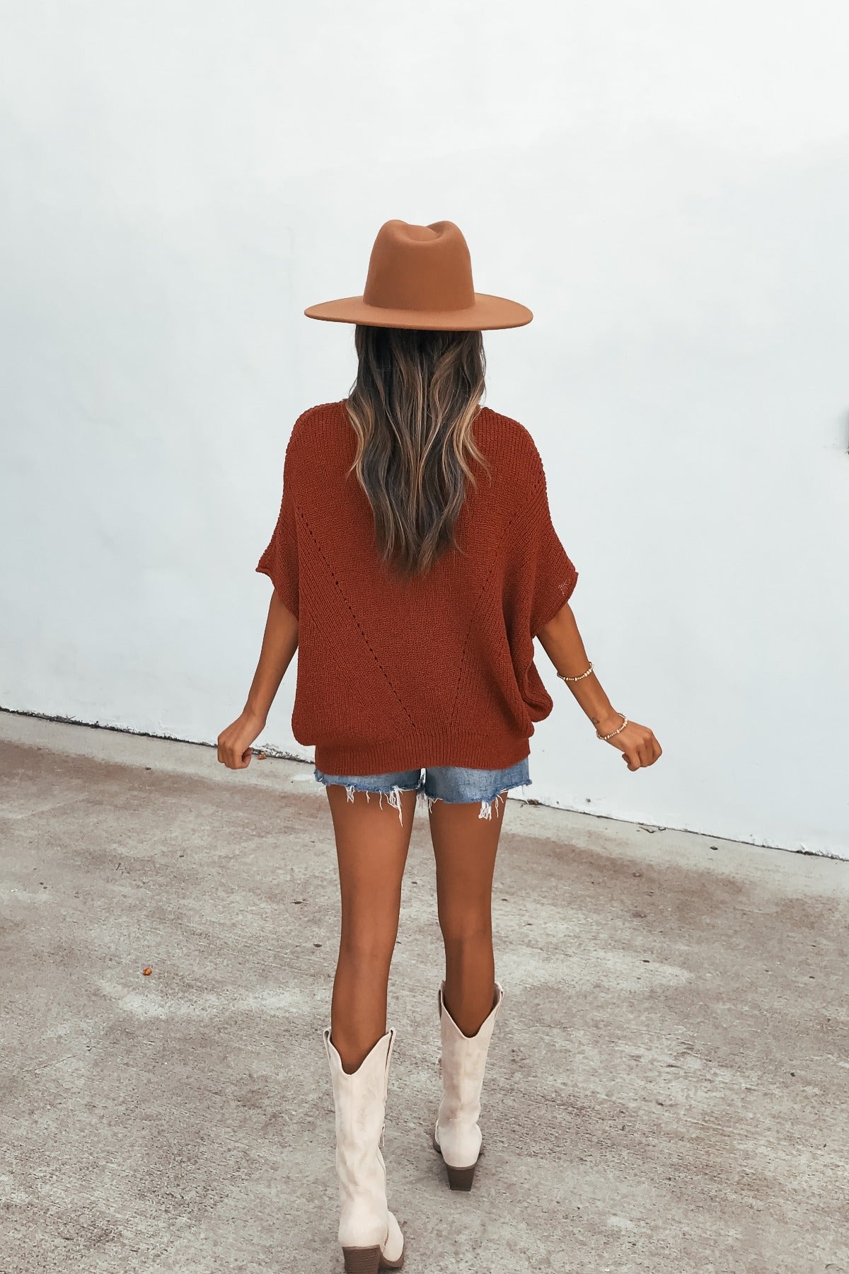 Woman in a brown hat and Cassia Boatneck Sweater - Rust pairs denim shorts with white boots, outdoors facing away from the camera.