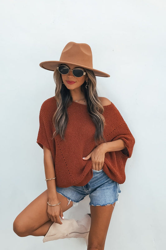 Woman in a brown hat, sunglasses, Cassia Boatneck Sweater - Rust, and denim shorts poses by a white wall, hand on hip and leg crossed.