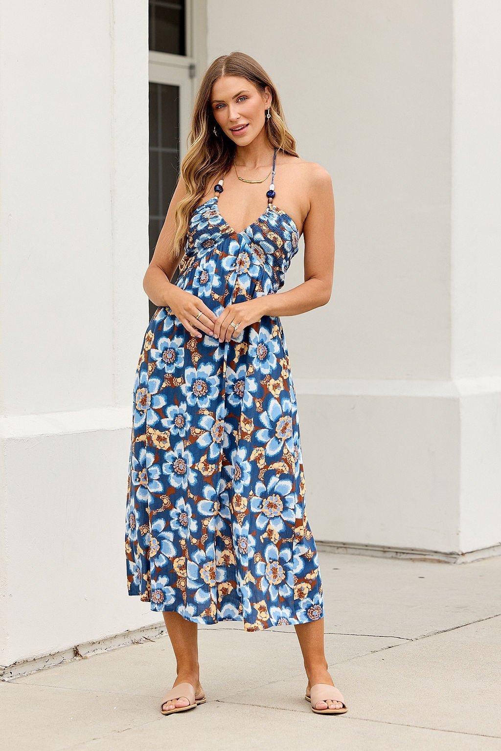 Woman wears Catalina Halter Midi Dress - Navy, stands outside a white building in sandals, smiling at the camera.