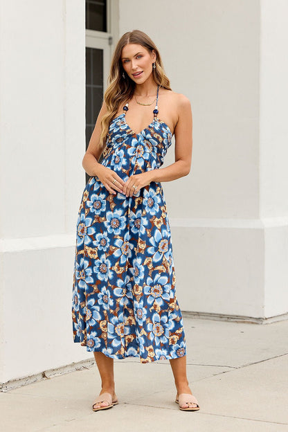 Woman wears Catalina Halter Midi Dress - Navy, stands outside a white building in sandals, smiling at the camera.