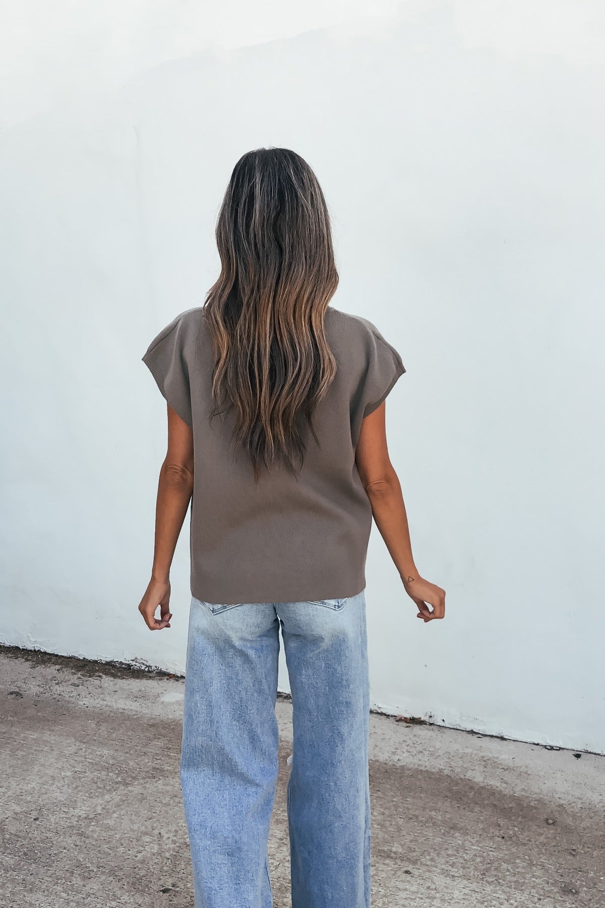 Woman with long wavy hair in a relaxed fit Catalina Striped Top - Olive and light blue jeans, standing facing a white wall.