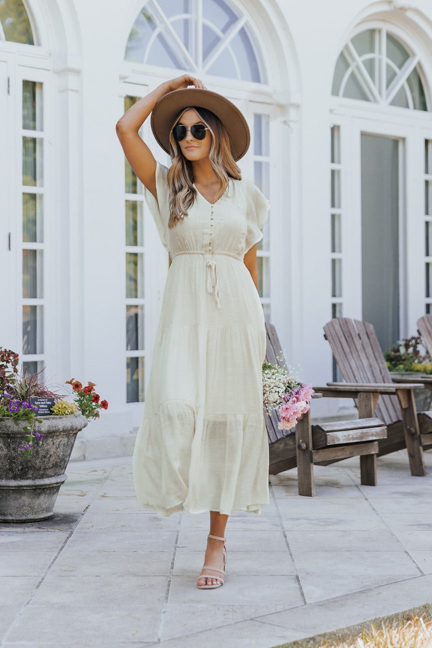 Woman in a Celine Crochet Lace Tiered Boho Midi Dress, hat, and sunglasses strolls on a patio with chairs, flowerpots, and arched windows.