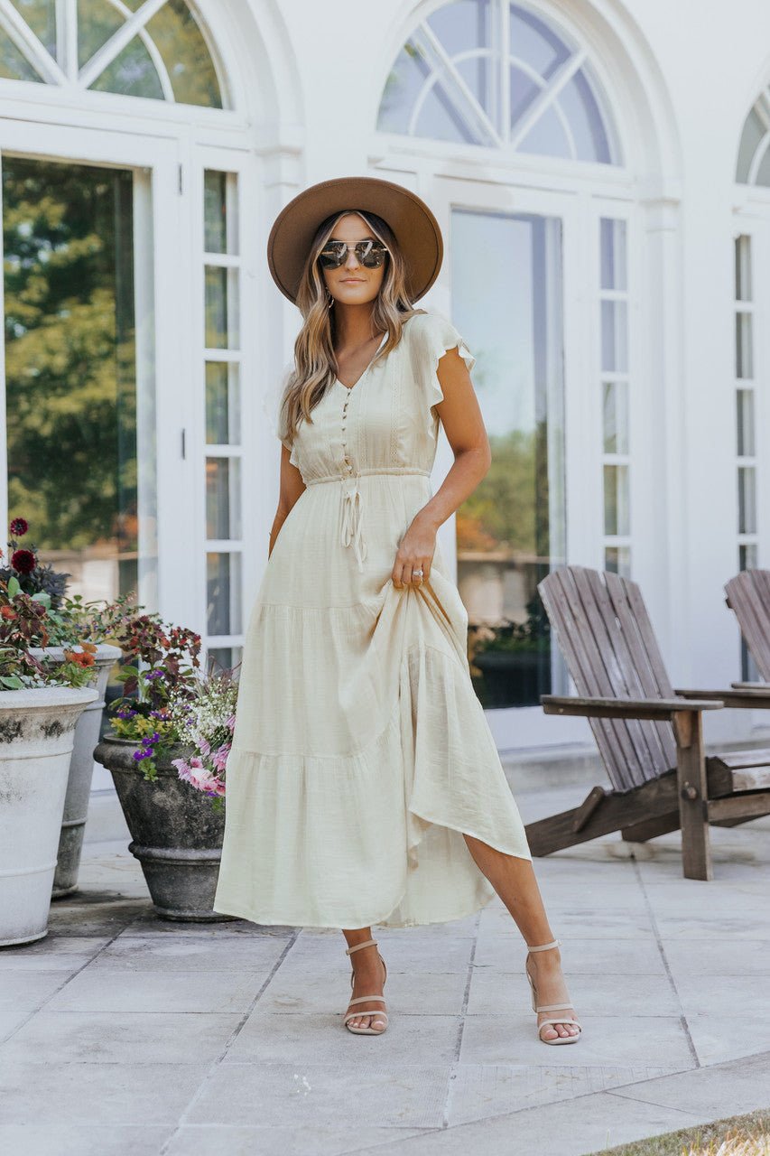 Woman in the Celine Crochet Lace Tiered Boho Midi Dress stands on a patio by flowers, wooden chairs, and large windows.