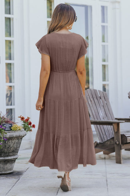 A woman in the Celine Crochet Lace Tiered Boho Midi Dress - Brown stands outside by wooden chairs and a planter with flowers.