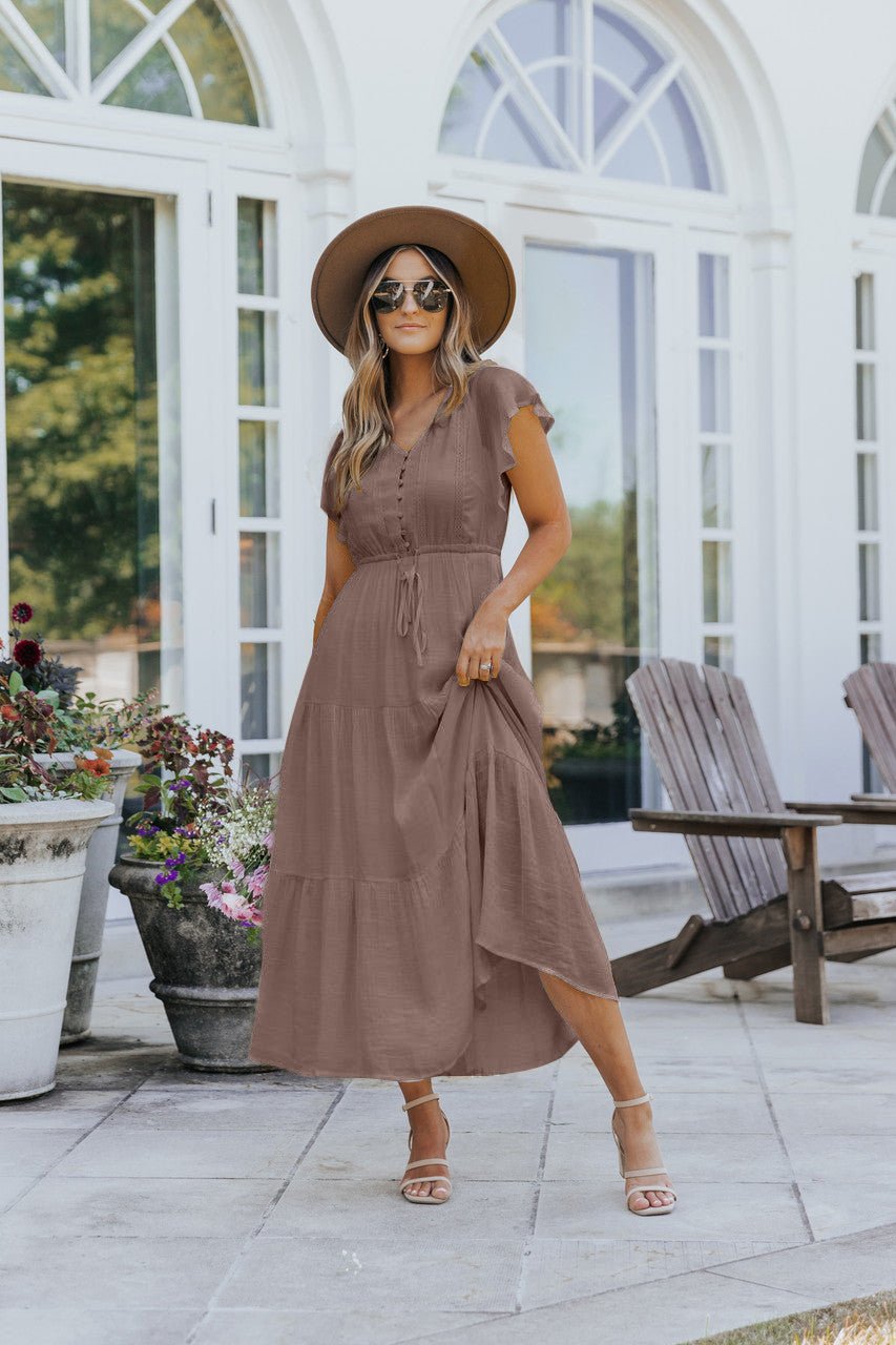 Woman in the Celine Crochet Lace Tiered Boho Midi Dress - Brown stands on a patio near wooden chairs and potted flowers.