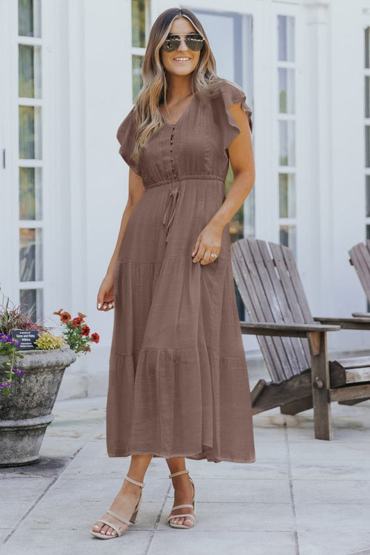 Woman in the Celine Crochet Lace Tiered Boho Midi Dress - Brown, standing outdoors near wooden chairs and a flower planter.