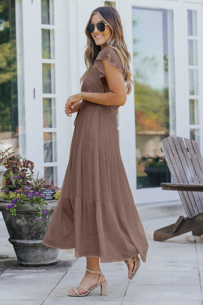 Woman in Celine Crochet Lace Tiered Boho Midi Dress - Brown, smiling on a patio with flowers, wooden chairs, and sandals.