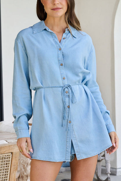 A woman wearing the Chambray Tie Button Down Romper, standing indoors.