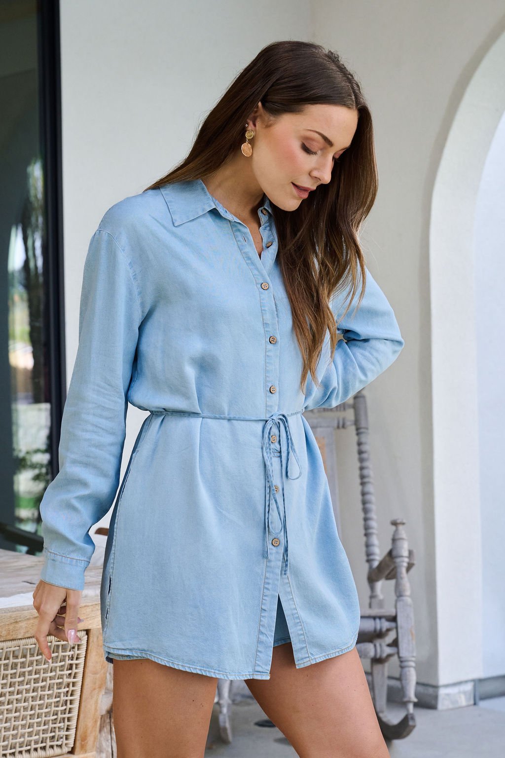 A woman wears the Chambray Tie Button Down Romper indoors, looking down with one hand on her hip.