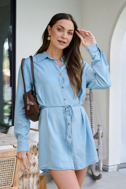A woman in a Chambray Tie Button Down Romper with a brown shoulder bag smiles outside by a table, touching her hair.