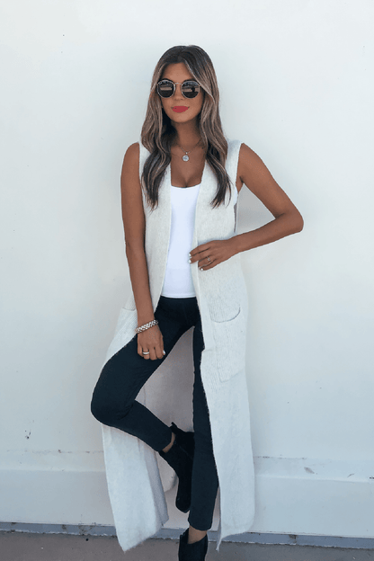 Woman in sunglasses and black pants models the Champagne Open Front Long Cardigan, a chic fall staple, against a white wall.