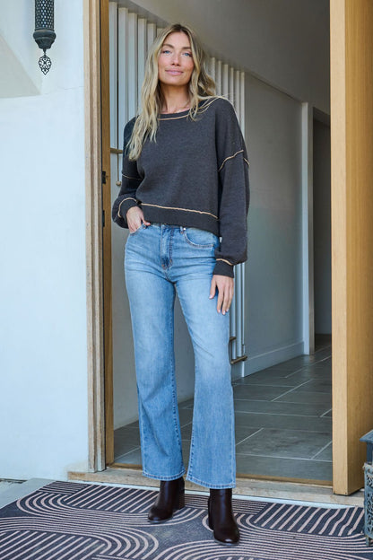 A woman smiles at the camera in a Charcoal Contrast Seam Detail Pullover, light blue jeans, and dark boots.