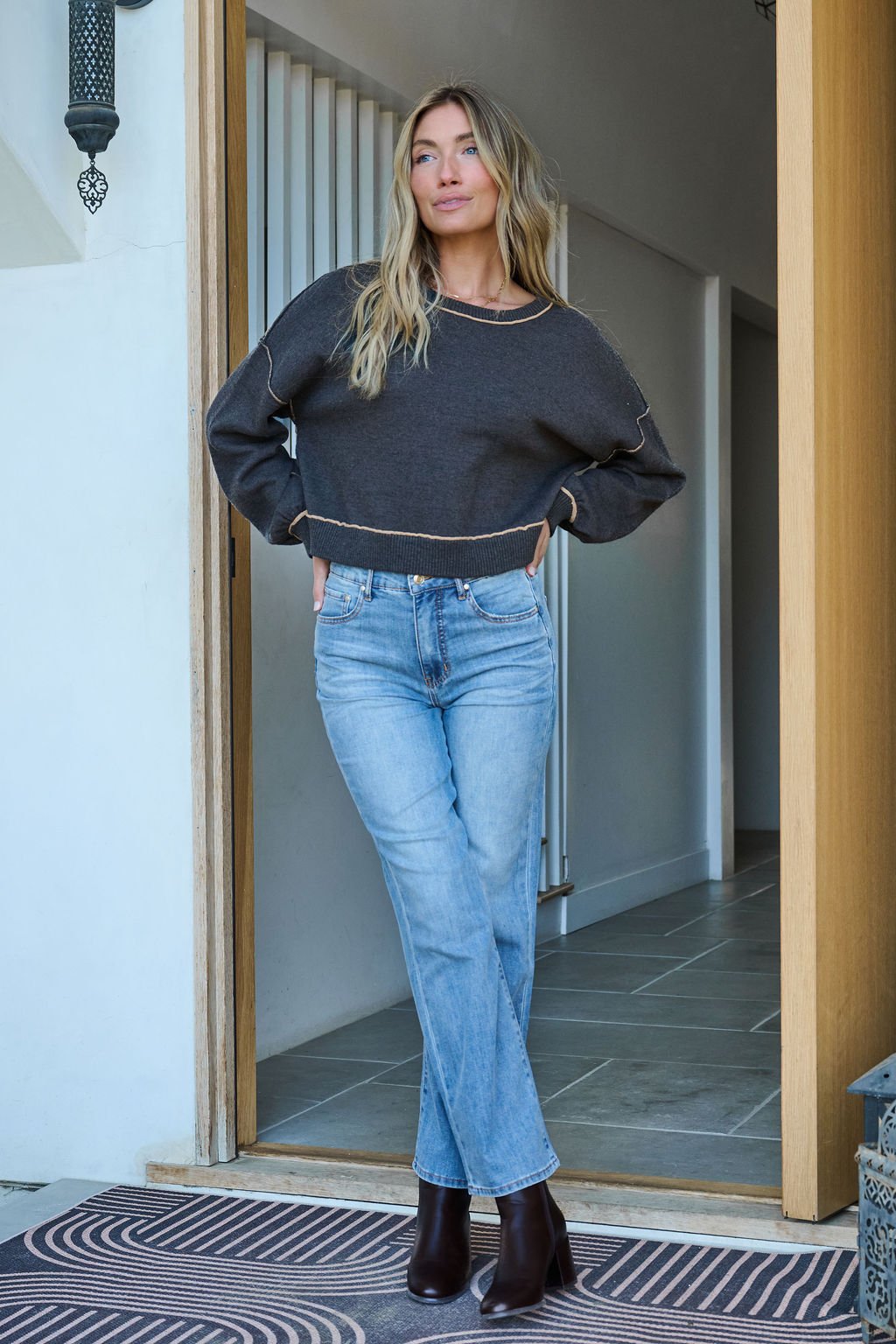 A woman stands in a doorway wearing a Charcoal Contrast Seam Detail Pullover, light blue jeans, and dark ankle boots, hands on hips.