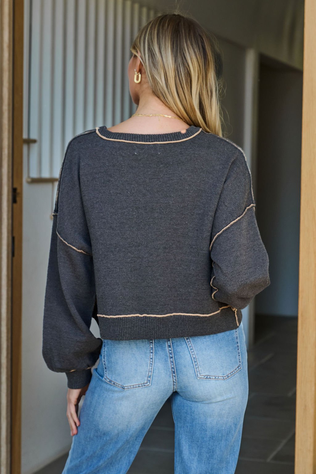 A woman with long blonde hair wears a Charcoal Contrast Seam Detail Pullover and light blue jeans, seen from behind indoors.